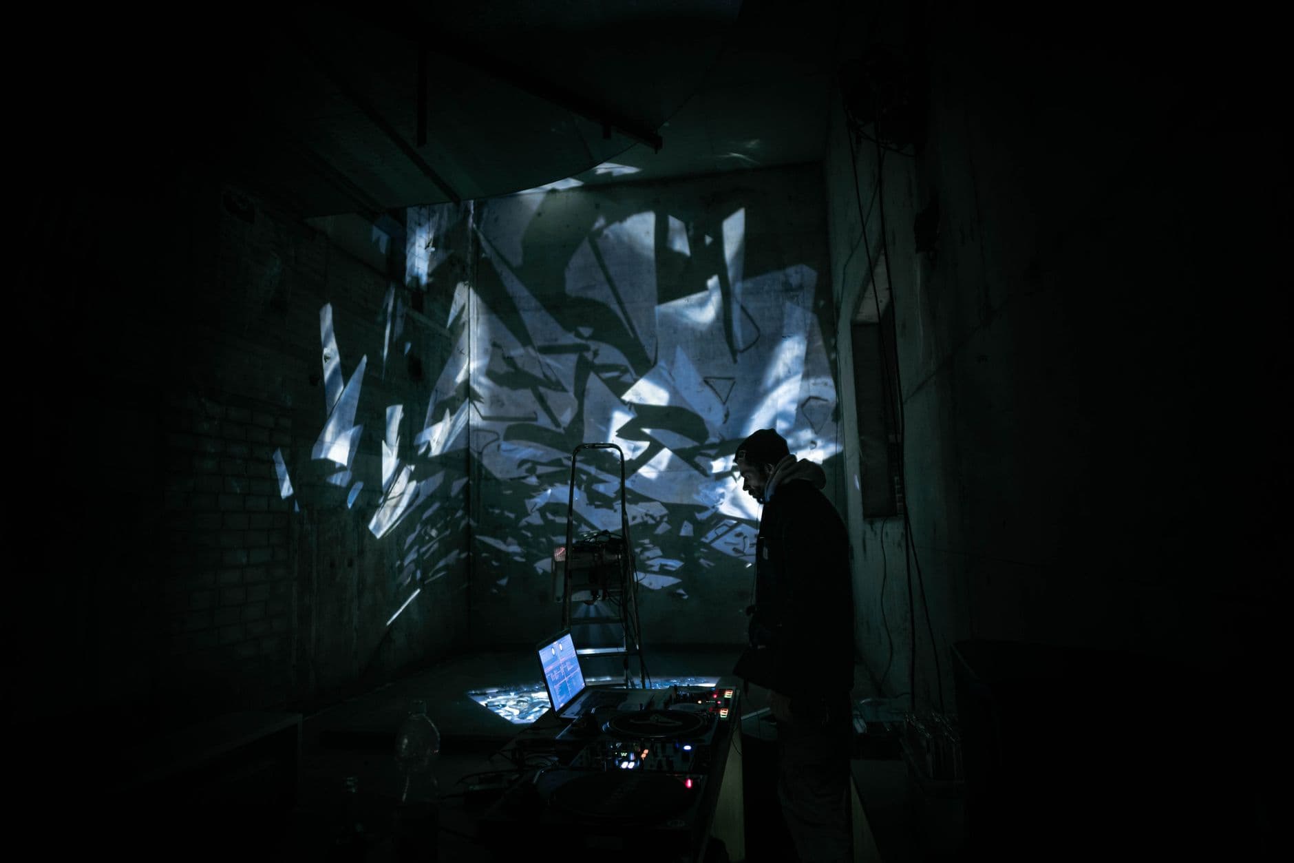 A DJ performing in a dark urban setting with abstract light projections creating a moody atmosphere.