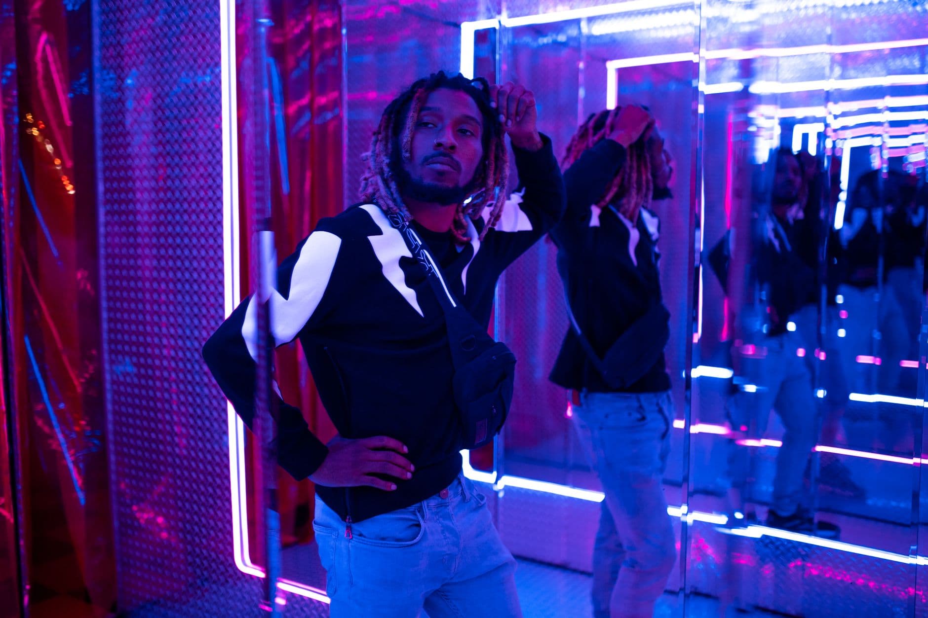 A stylish man poses in a neon-lit room with vibrant mirrors, creating an artistic effect.